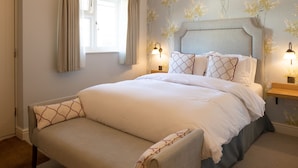 A neatly made bed with a headboard, a bedside table with a lamp, and a bench with patterned cushions. - The Inn West End (Woking)
