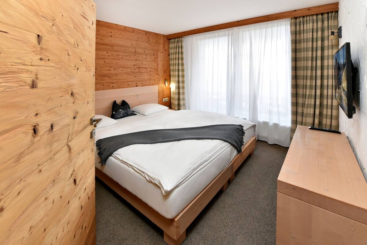 A bedroom with a wooden headboard, a bed with white bedding, a bedside table, a TV mounted on the wall, and a window with curtains.