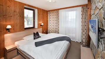 A bedroom with a wooden headboard, a bed with white bedding, a nightstand, a lamp, a window with blinds, and a stone wall with a poster.