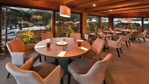 A dining area with round tables and cushioned chairs, large windows with a view of a snowy landscape, and a stone wall with a Christmas tree. - APLEND Hotel Kukučka a Rezidencie (Vysoké Tatry)
