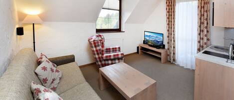 A cozy living room with a sofa, a plaid armchair, a wooden coffee table, a TV, and a kitchen area with a sink.