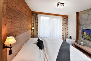 Ett hotellrum med en sänggavel i trä, en säng med vita sängkläder, ett litet nattduksbord med en lampa, ett mindre vitt nattduksbord och en vägg av sten med ett tv-apparat.