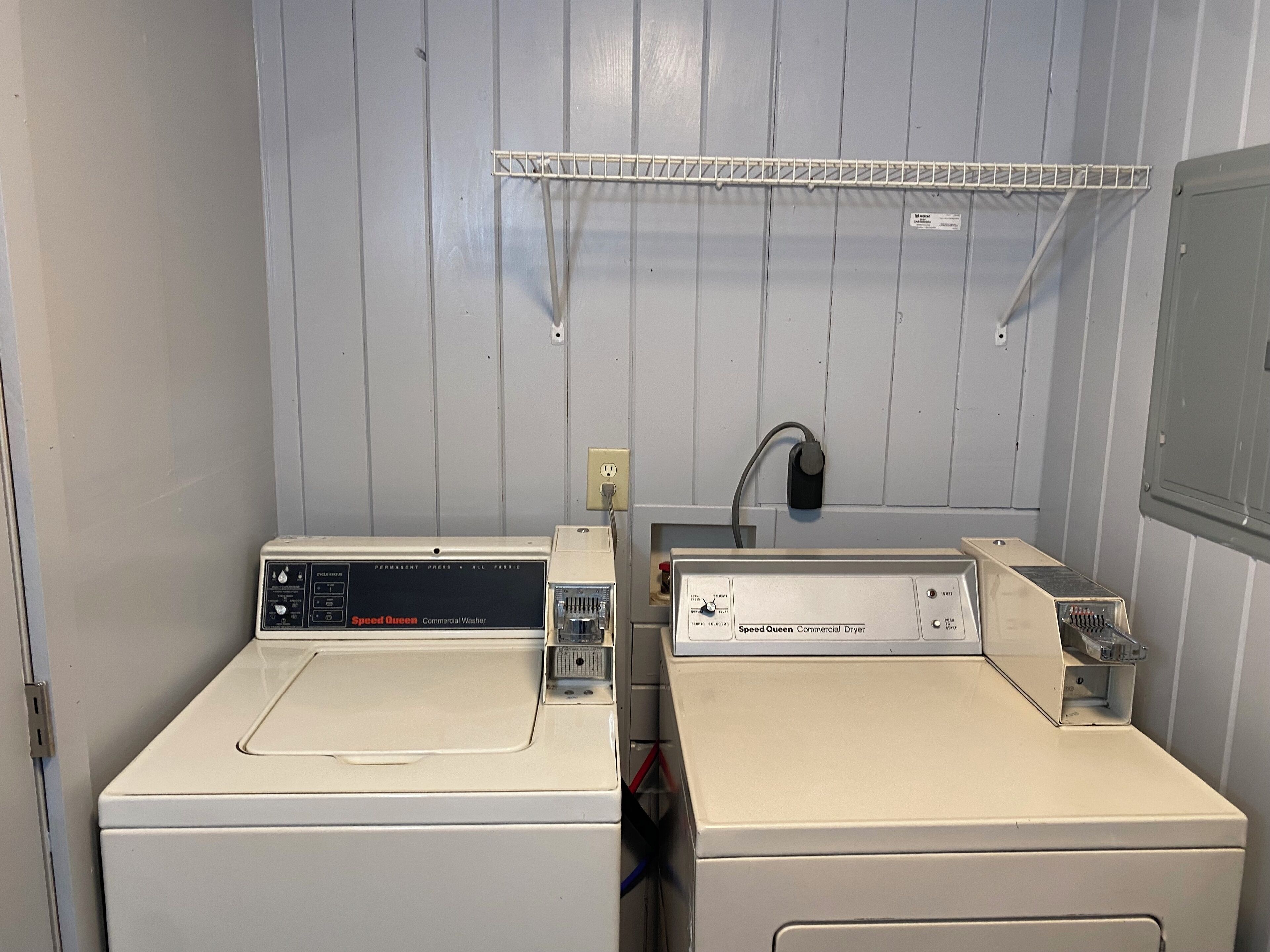 A laundry room with two white commercial washers and dryers, a metal shelf above, and a wall-mounted electrical outlet.