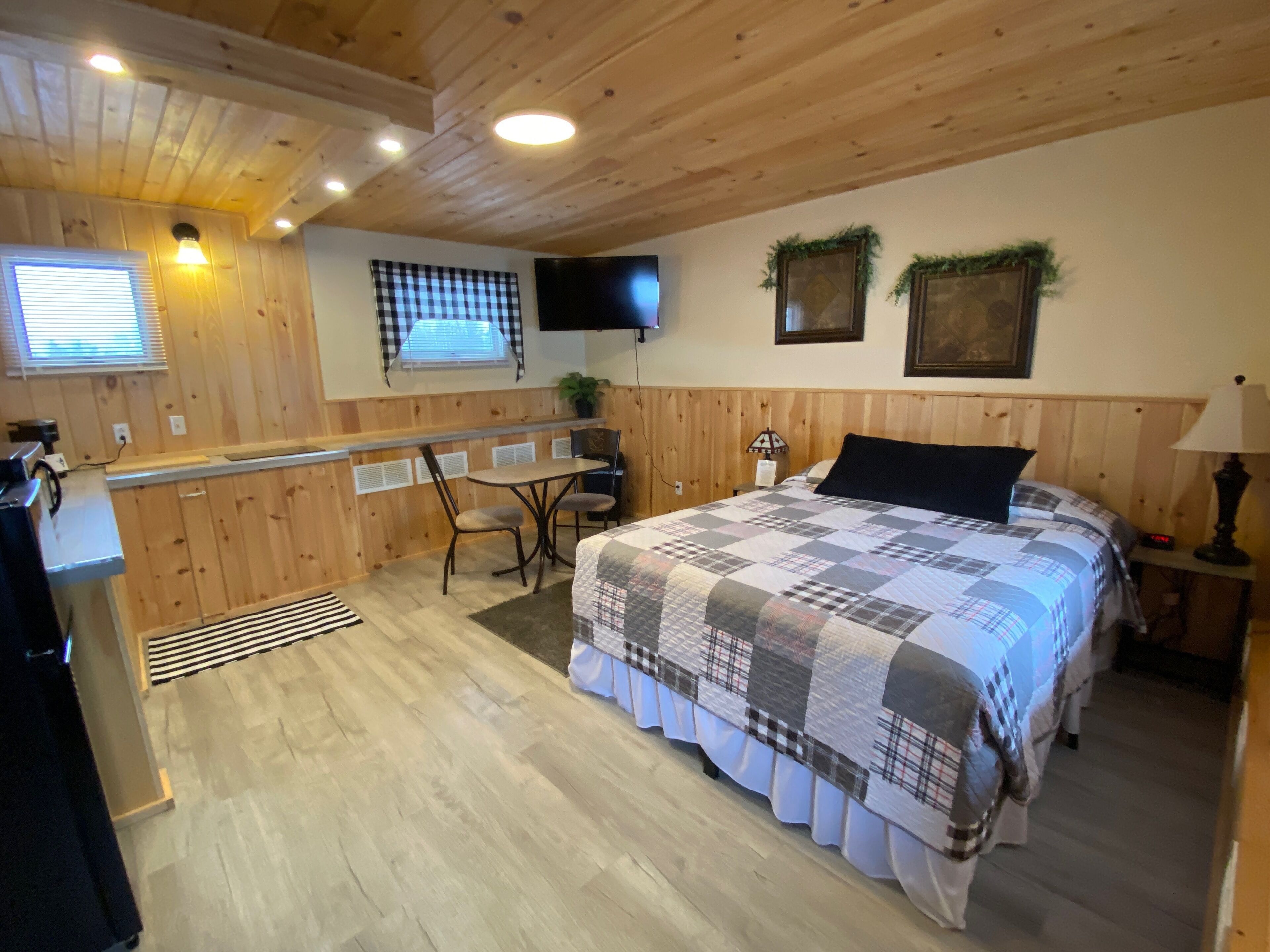 A cozy room with a bed, a desk, and a chair. There is a window with blinds, a flat-screen TV mounted on the wall, and a small table with a lamp.