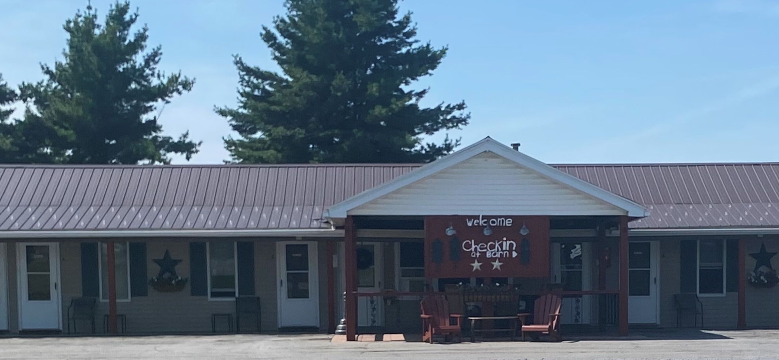 Een motel met een 'Welcome to Checkin Inn' bord, een overdekte veranda met stoelen, en een metalen dak.