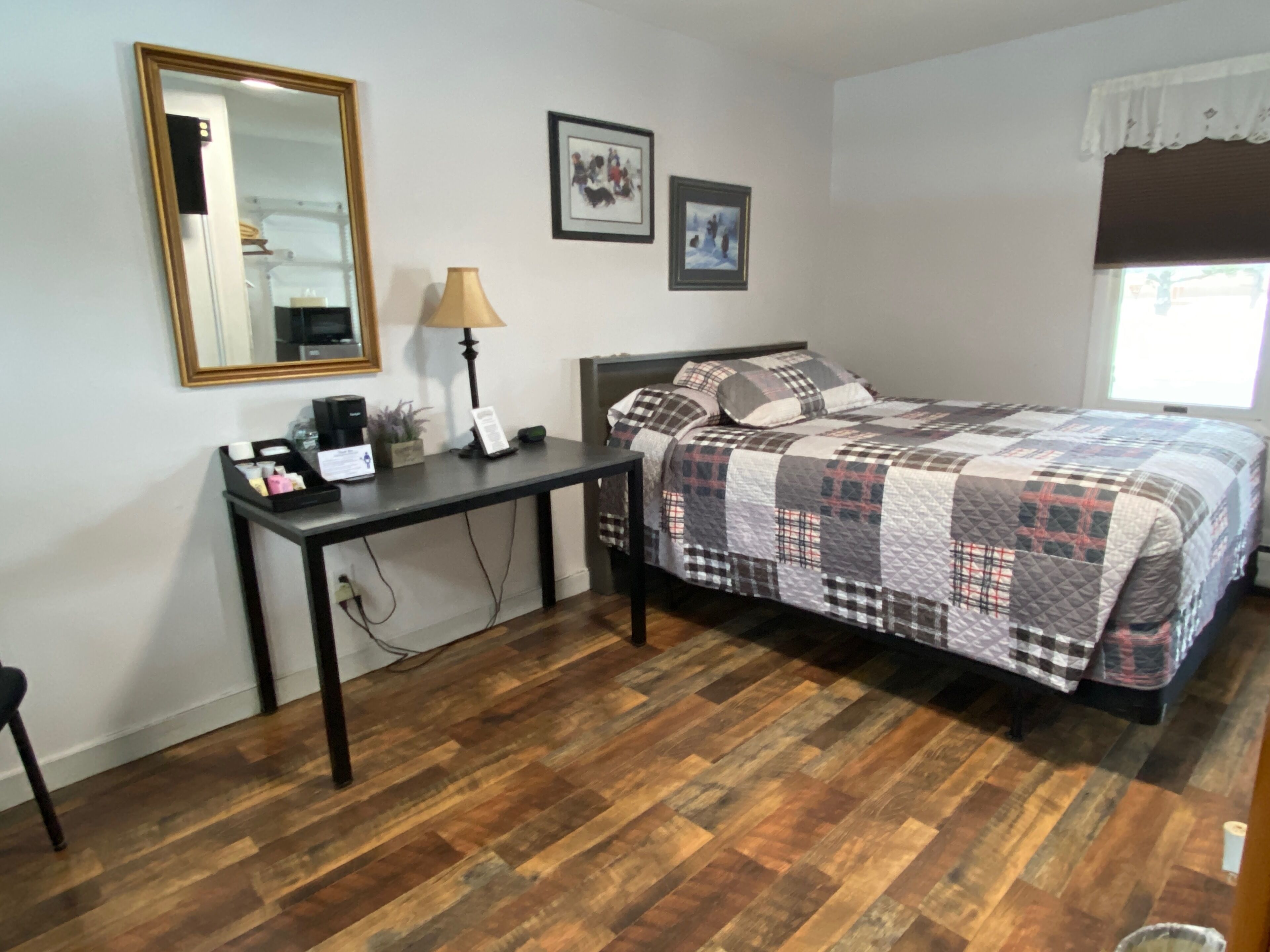A bedroom with a wooden floor, a bed with a checkered quilt, a desk with a lamp, and a mirror.