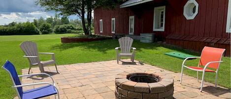 Un patio con un hogar para fogata, tres sillas Adirondack y un granero rojo al fondo.