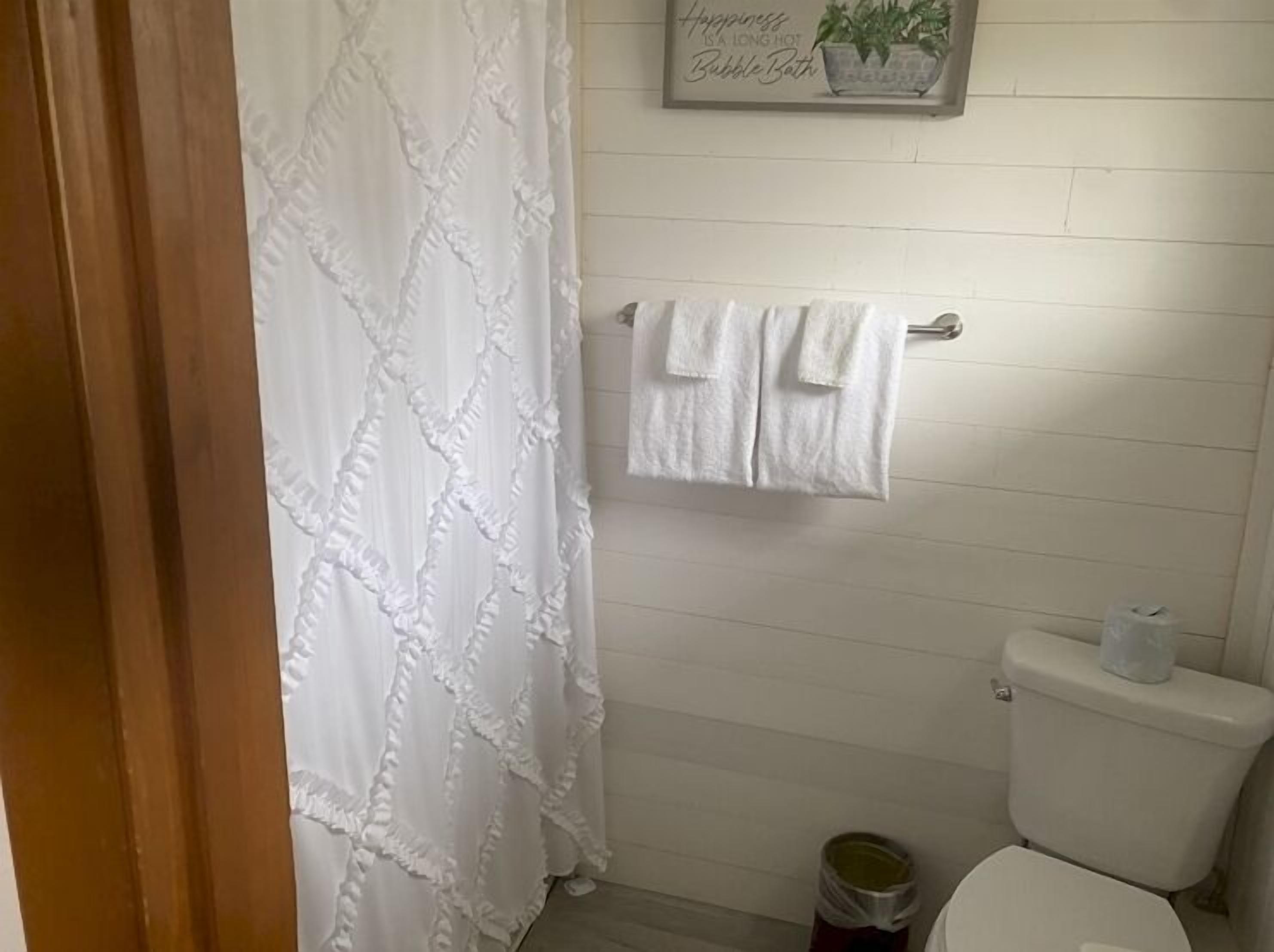 Un baño con cortina de ducha blanca, un perchero con dos toallas, un inodoro y un bote de basura.