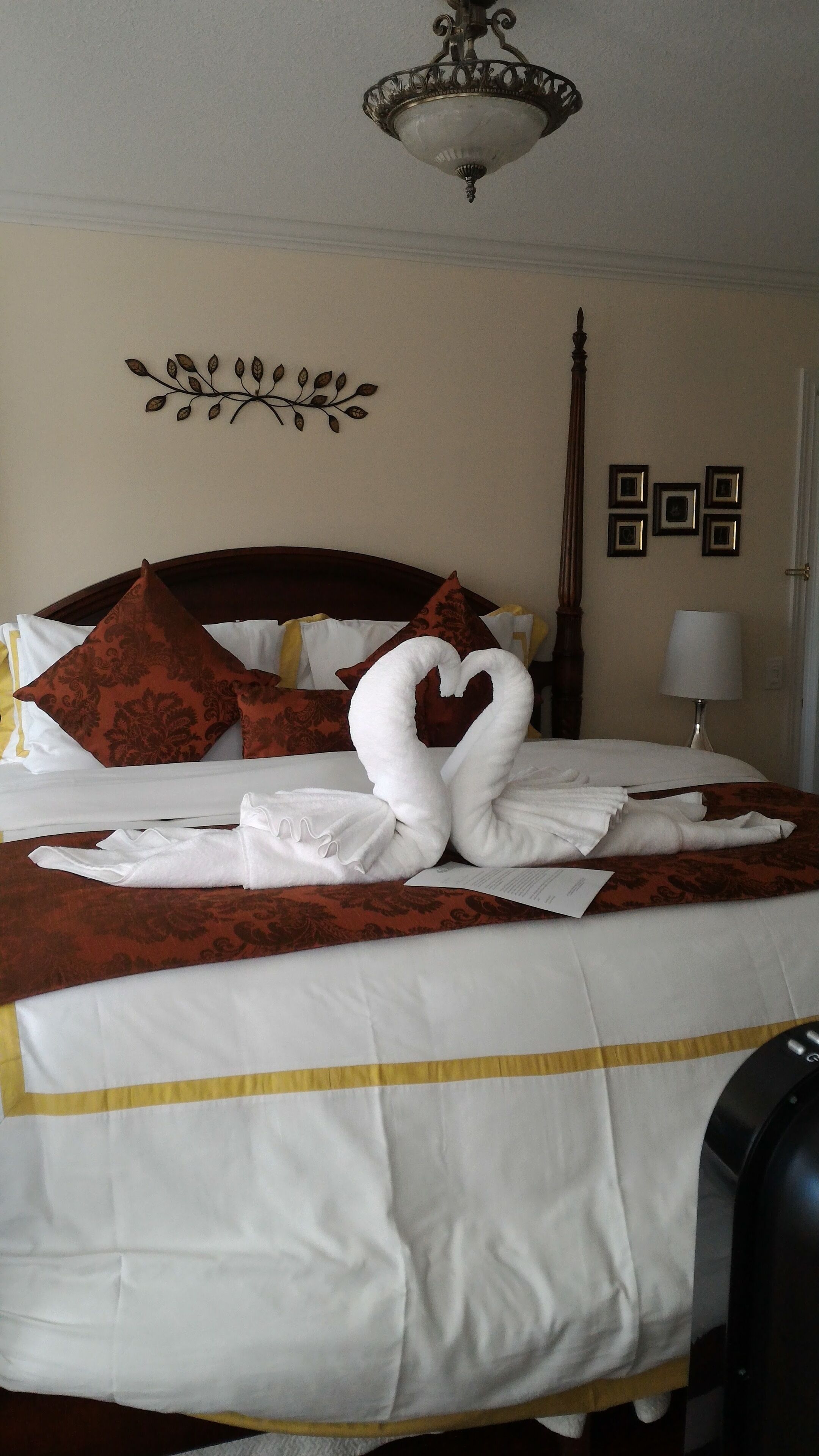 A neatly made bed with a heart-shaped towel arrangement, a decorative wall piece, and a lamp.