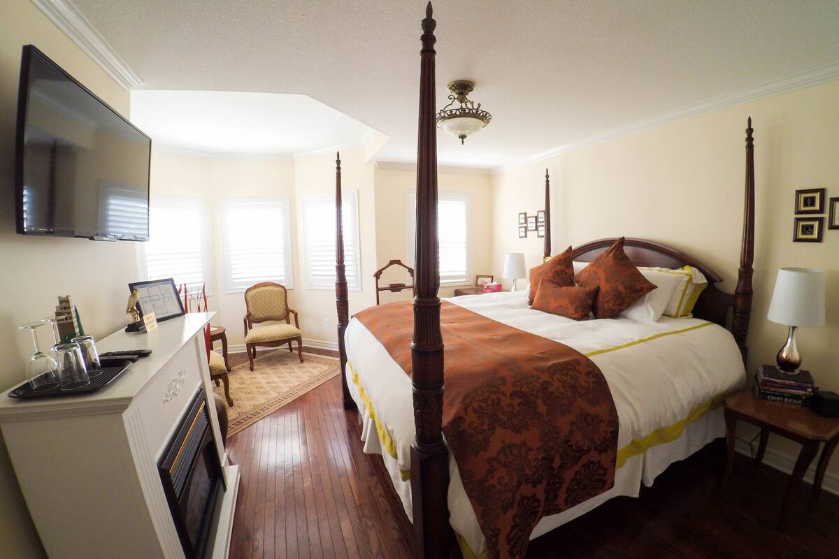 A four-poster bed with a wooden headboard, a bedside table with a lamp, a chair, and a flat-screen TV mounted on the wall.