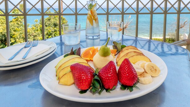 A table with a plate of fruit, a glass of water, and a glass of iced tea, set against a scenic ocean view. - Sunset Hotel (North Kynouria)