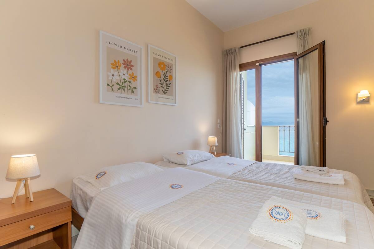 A hotel room with two beds, a nightstand, a lamp, and a window with a view of the sea.
