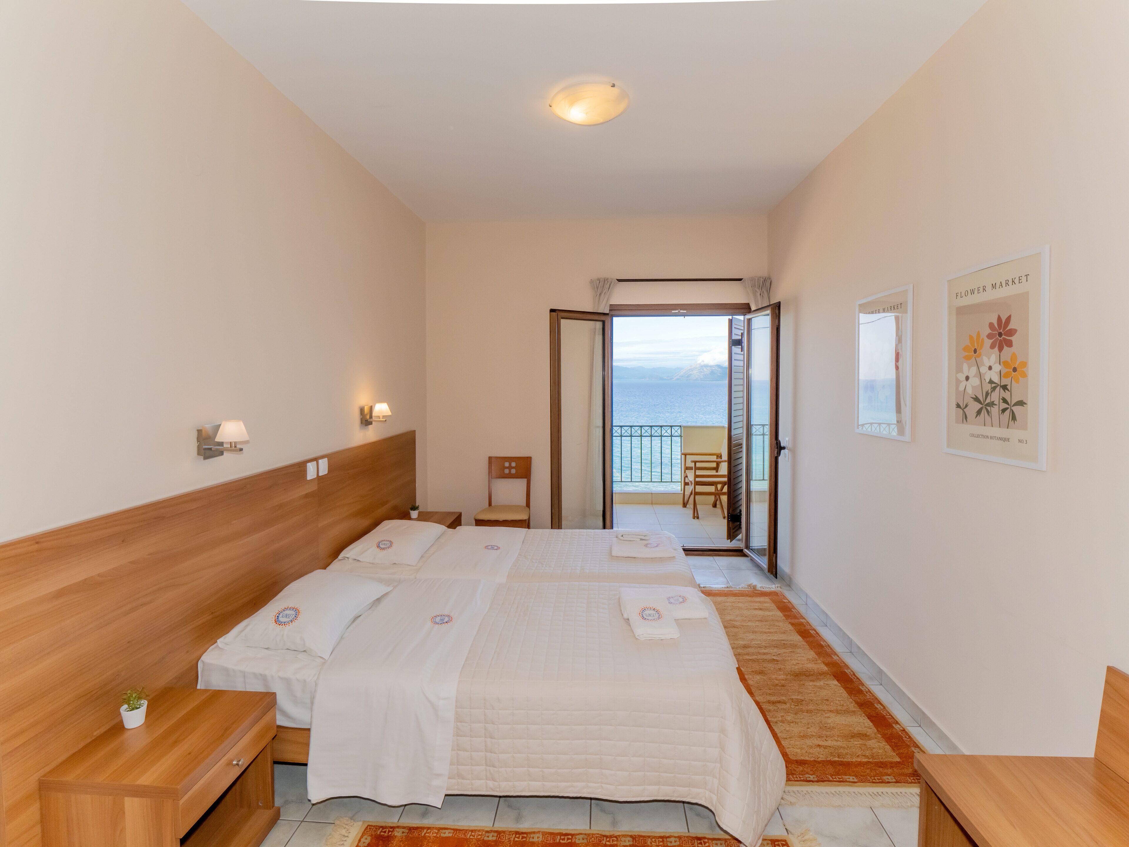 Une chambre d’hôtel avec deux lits, une tête de lit en bois, une petite table avec une plante et une vue sur la mer par une porte ouverte.