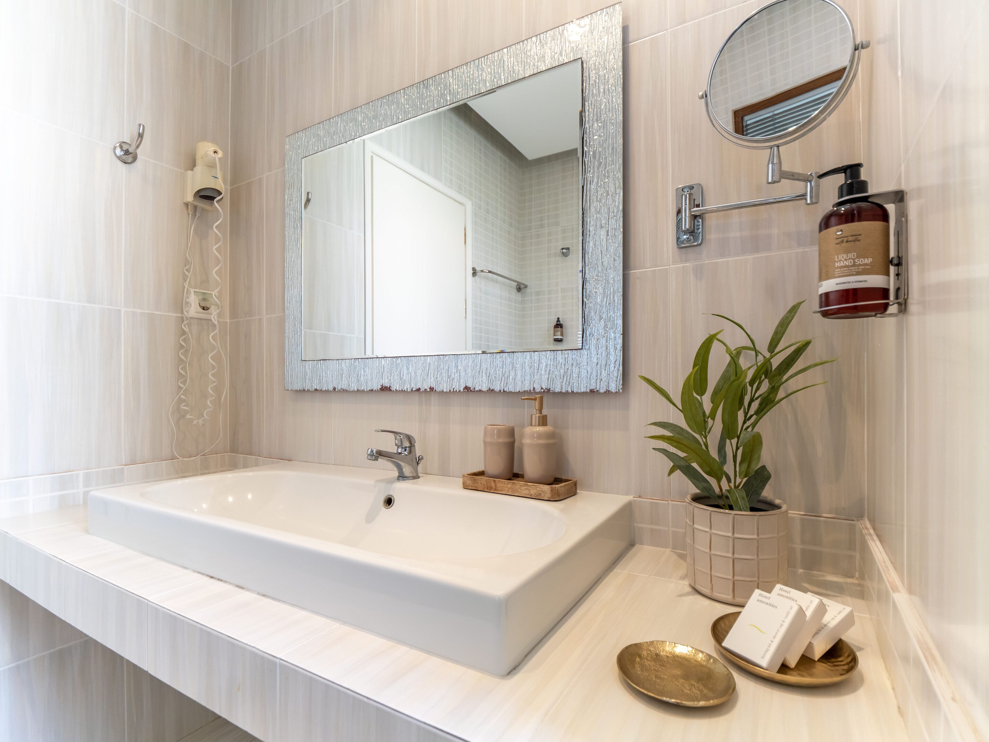Een moderne badkamer met een groot wit wastafel, een spiegel met decoratieve lijst, een handdouche en een kamerplant.