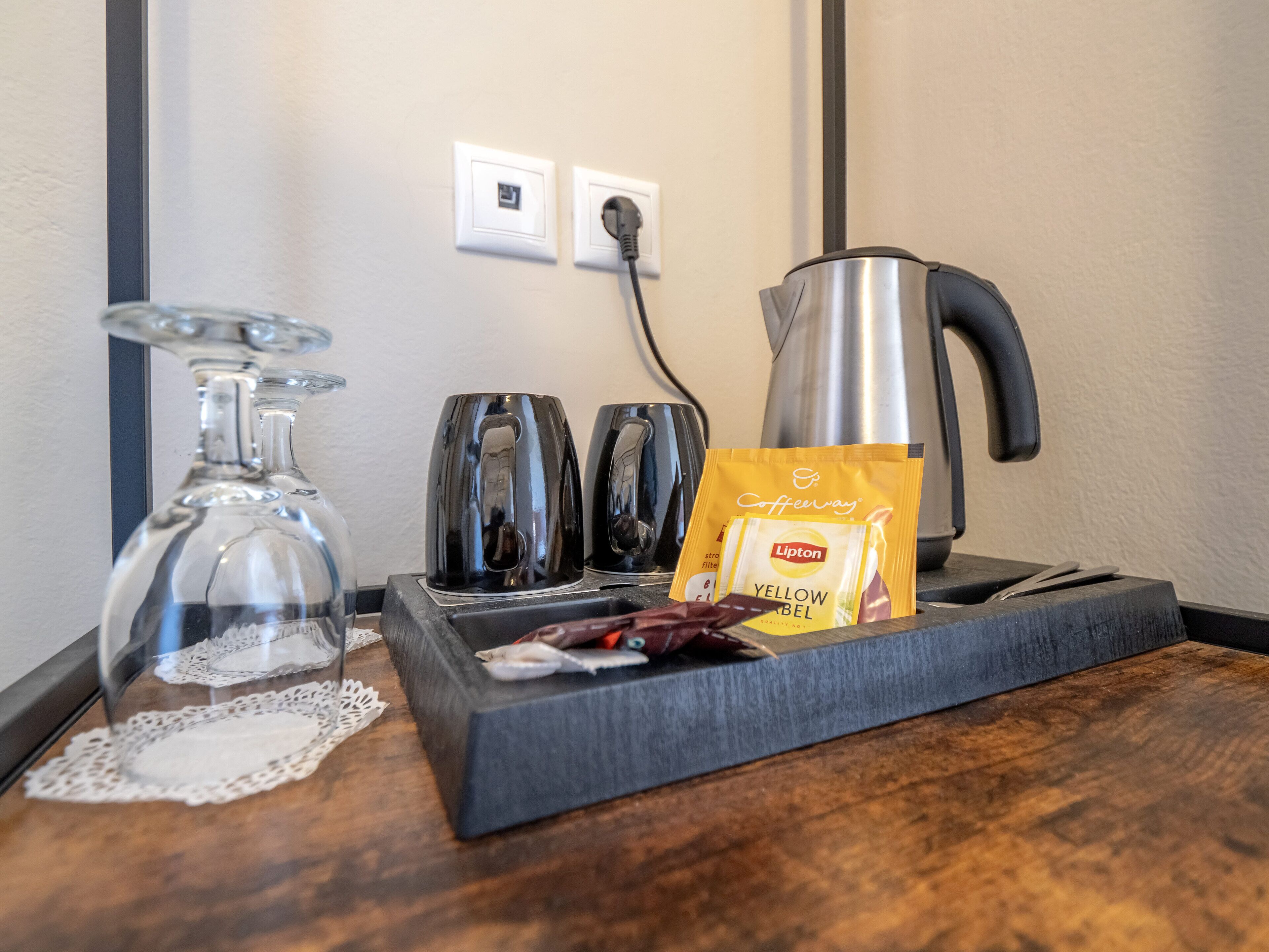 A coffee maker, two cups, a glass, a tea bag, and a power outlet on a wooden table.