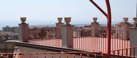 Una terraza en la azotea con barandillas rojas y balaustradas de piedra, con vistas a la ciudad y al mar.
