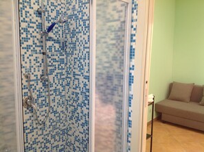 Une douche avec un mur recouvert de mosaïques bleues, un canapé beige et une petite table avec une lampe dans l’espace de vie.