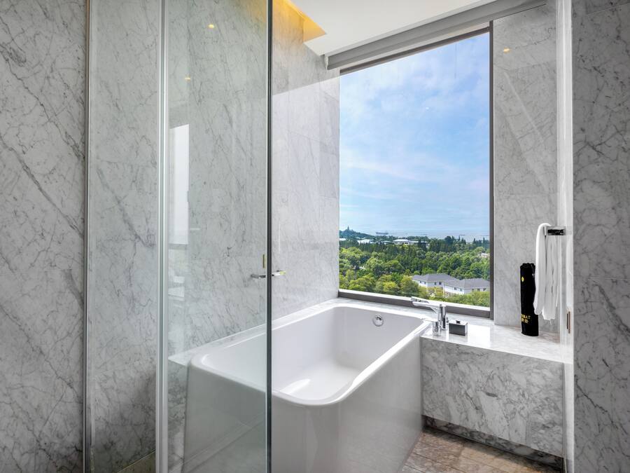 A modern bathroom with a glass-enclosed shower, a freestanding bathtub, and a window offering a city view.