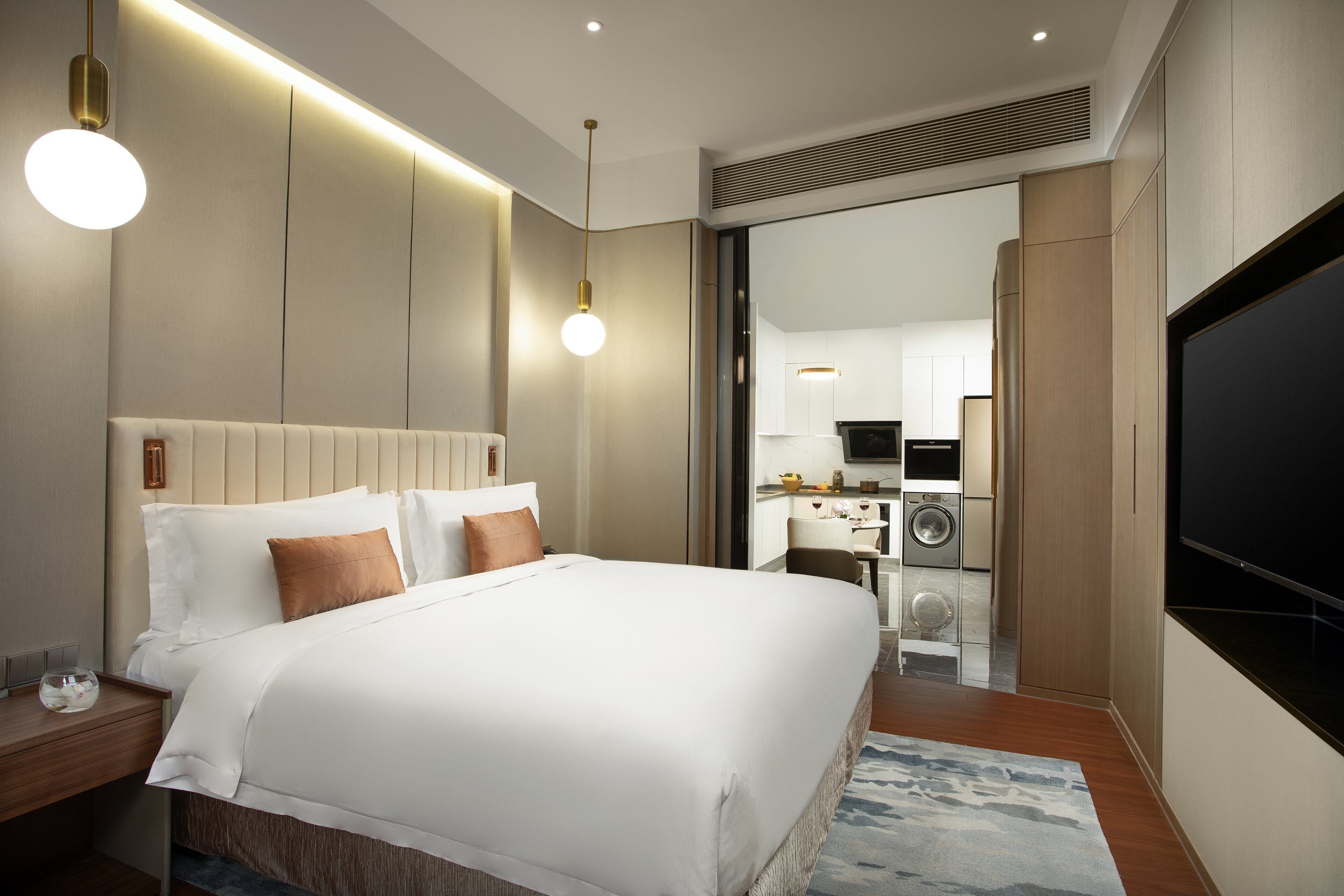 A modern hotel room with a large bed, bedside tables, a flat-screen TV, and a kitchenette area.