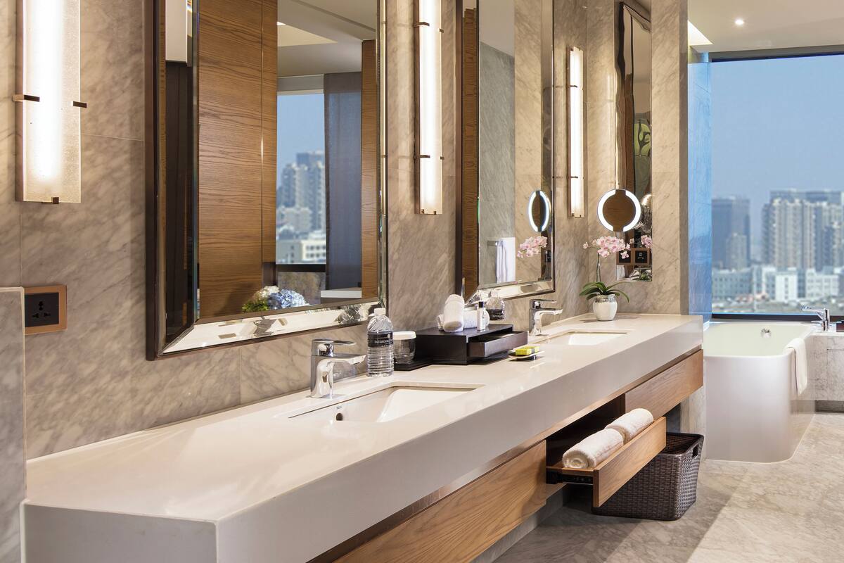 Een moderne badkamer met een grote spiegel, twee wastafels, een bad en uitzicht op de stad.