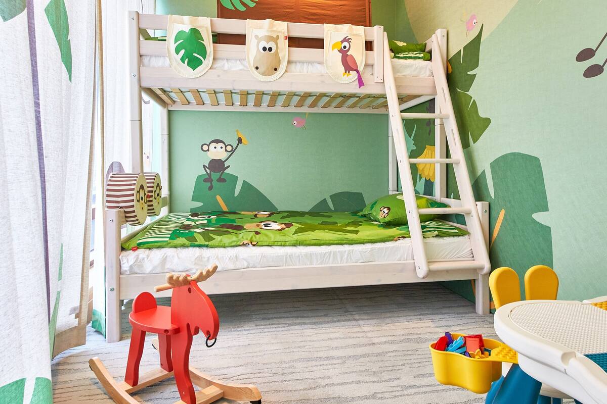 Een stapelbed met een groene blad- en dierenwanddecoratie, een klein rood schommelpaard en een gele opbergbox voor speelgoed.