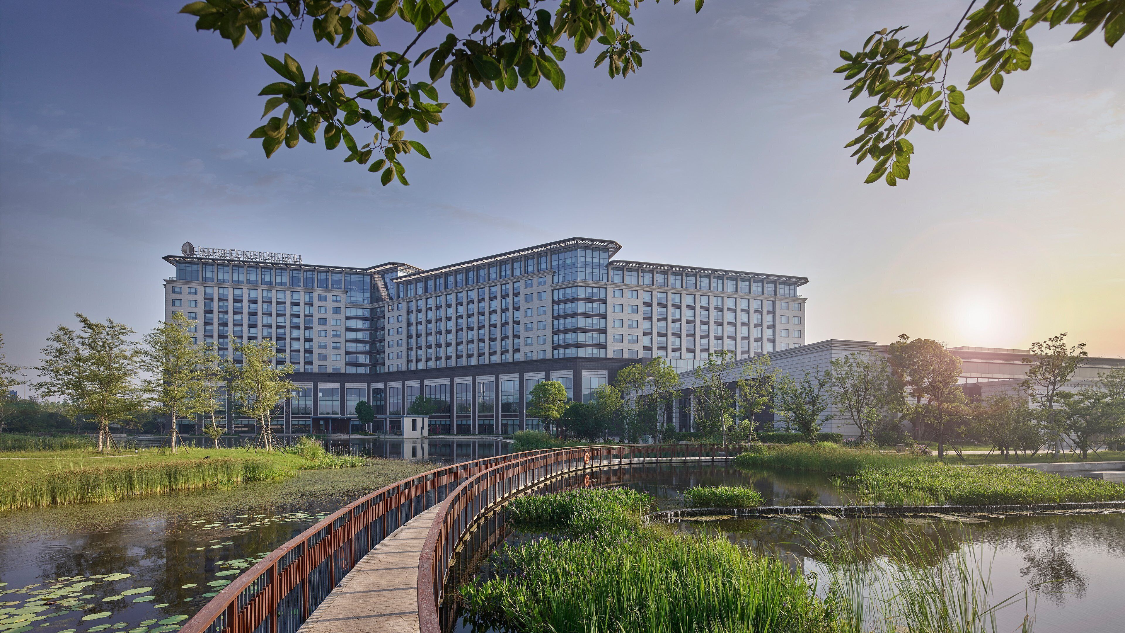 Un hôtel moderne doté d’une grande façade en verre, d’un pont enjambant un étang et entouré de verdure.