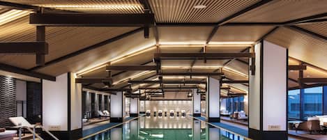 A modern indoor swimming pool with a reflective floor, surrounded by lounge areas and a high ceiling with wooden slats.