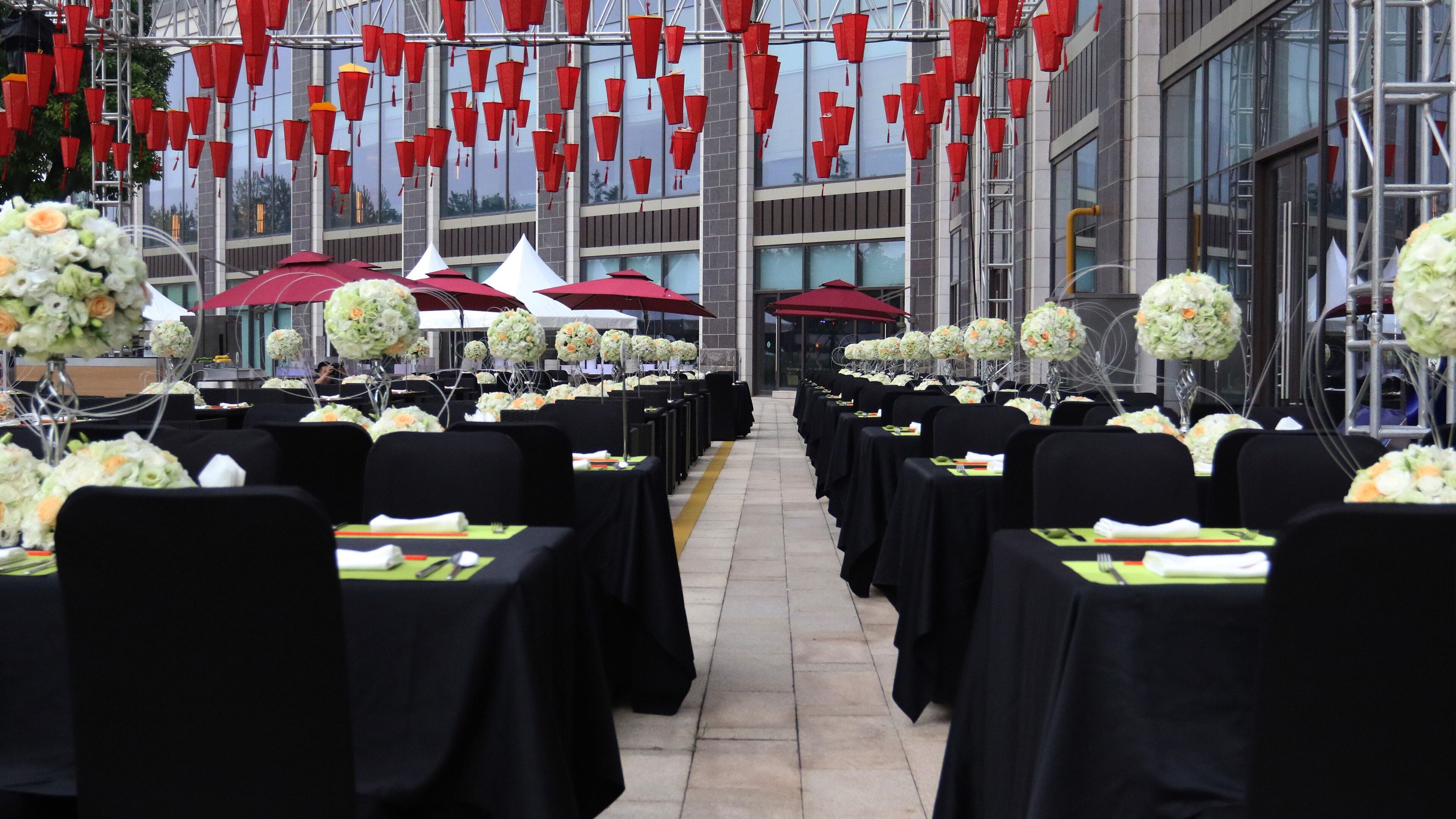 A banquet hall with rows of tables set for a formal event, featuring white floral centerpieces and red umbrellas.