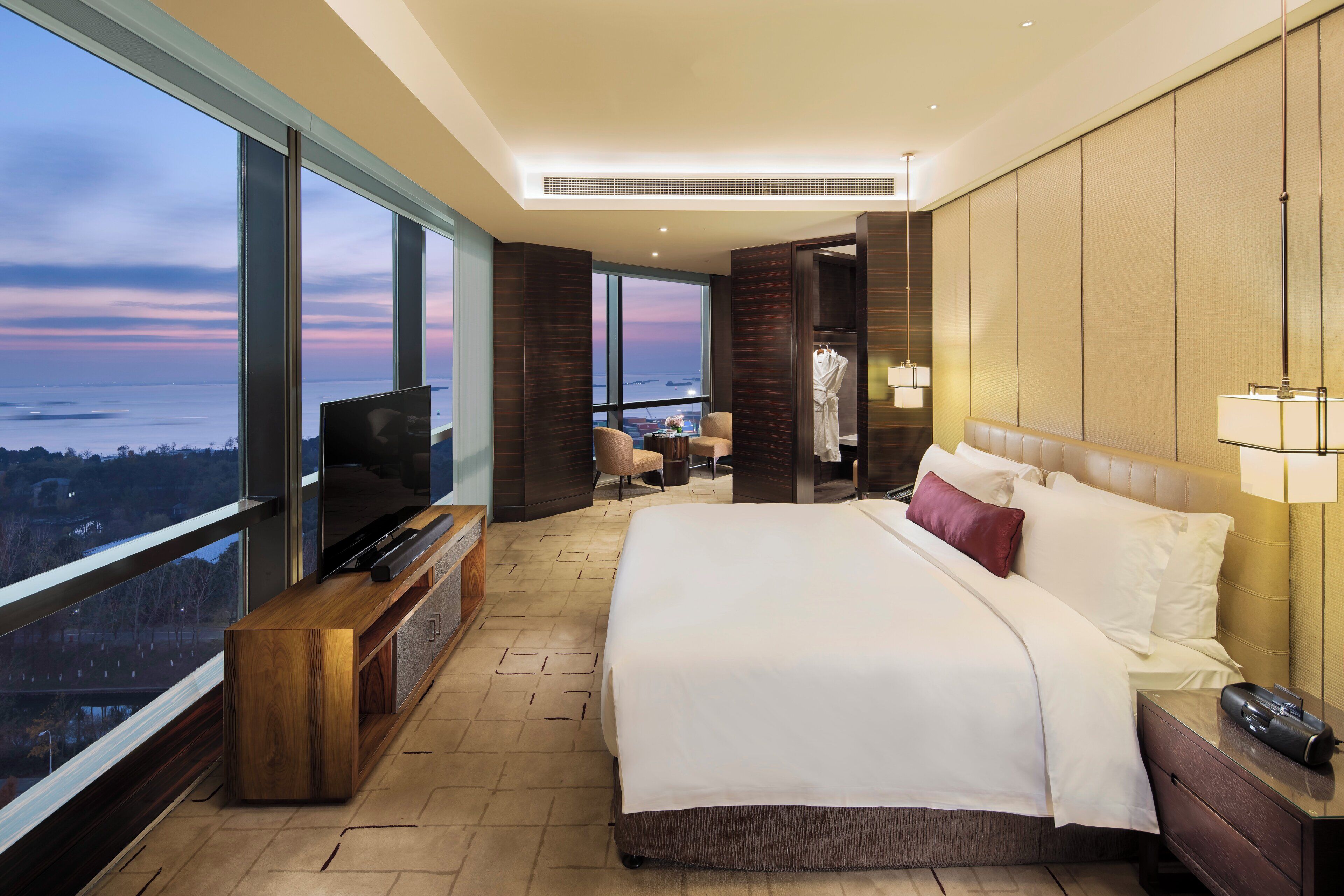 A modern hotel room with a large bed, a TV, a desk, and a view of the cityscape.