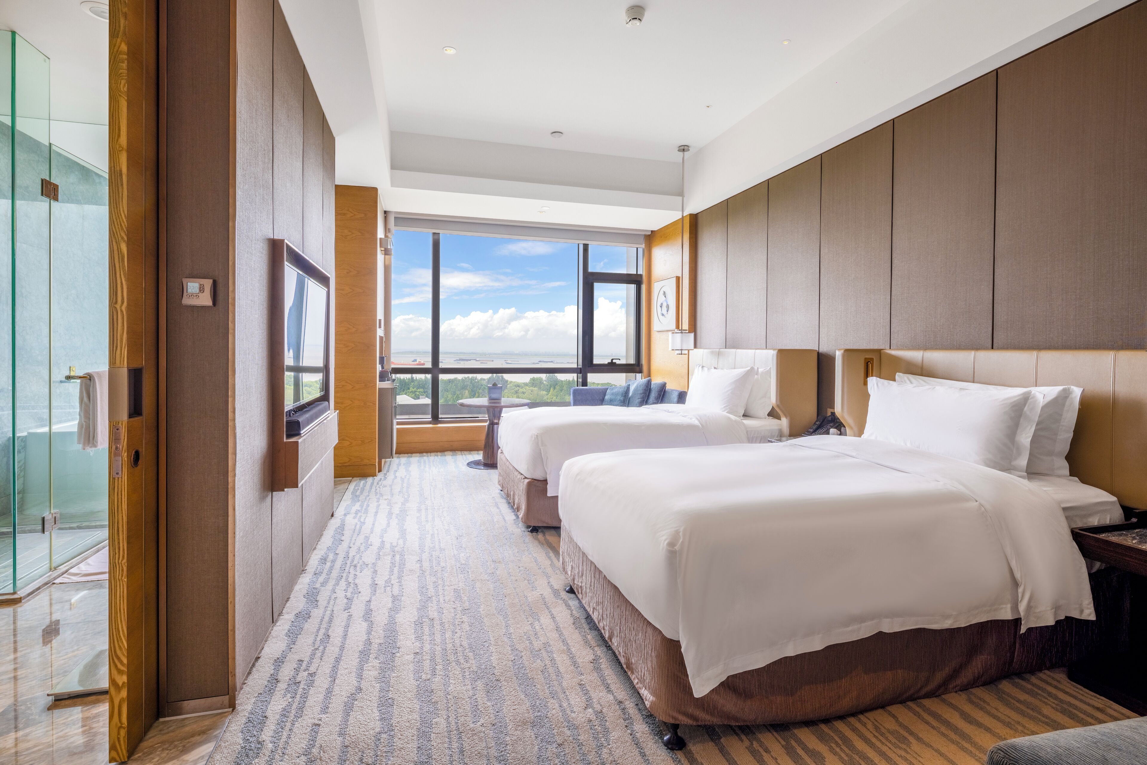 A hotel room with two beds, a large window with a city view, a glass shower enclosure, and a wooden panel wall.