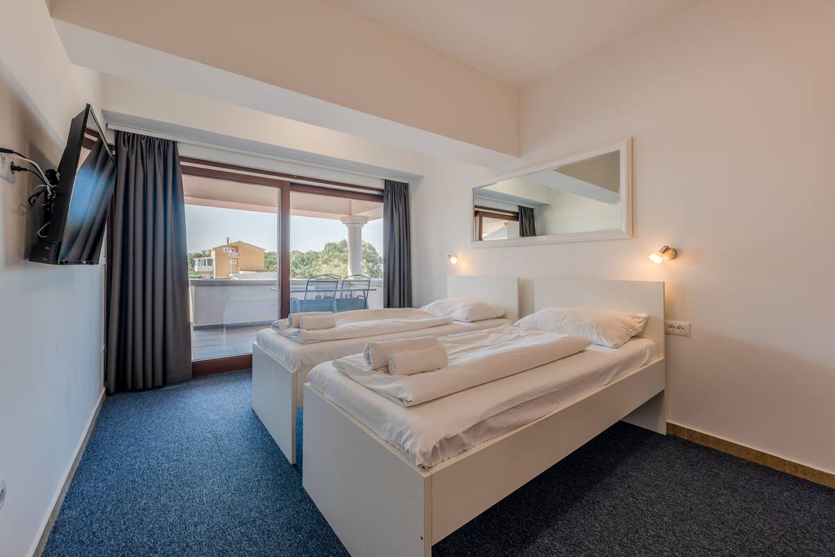 A hotel room with two beds, a large window with curtains, a flat-screen TV mounted on the wall, and a small table with a lamp.