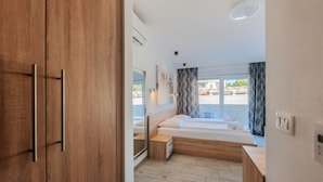 Une chambre à coucher moderne avec une armoire en bois, un lit recouvert de linge de maison blanc, une table de chevet et une vue sur l’extérieur.