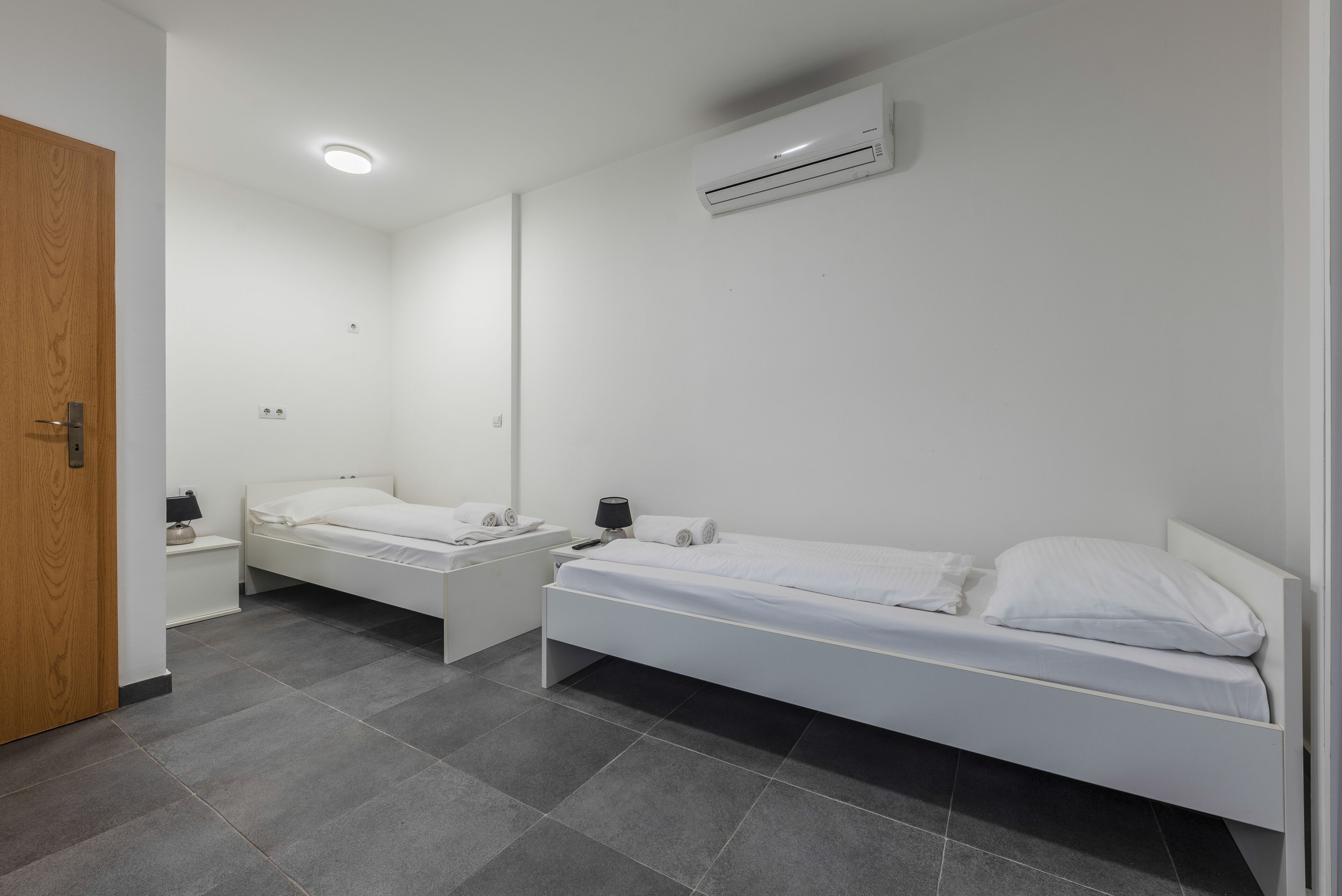 Camera da letto con due letti singoli, una porta in legno, un comodino bianco con una lampada e un condizionatore d'aria a parete.