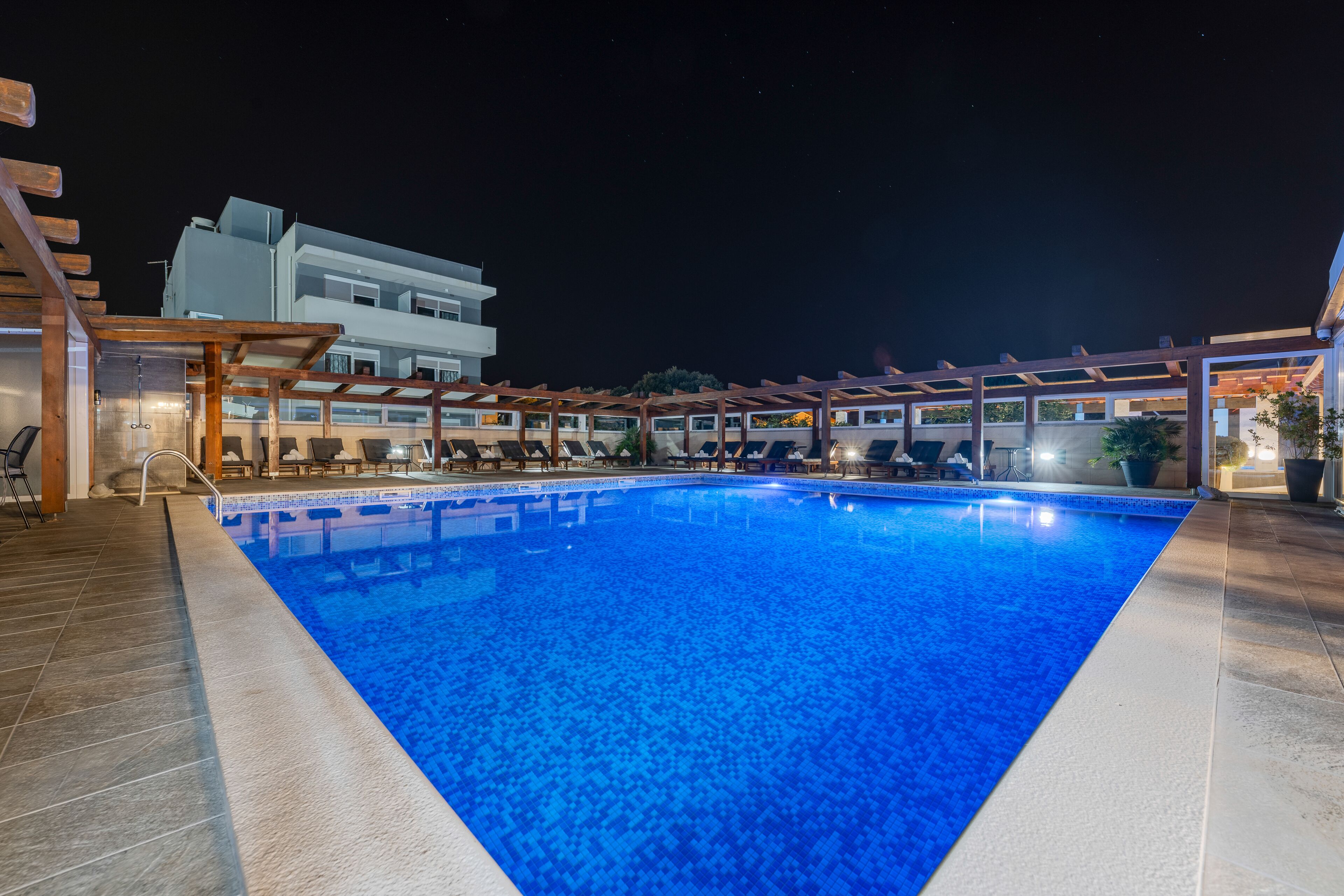A swimming pool with a wooden deck and lounge chairs, surrounded by a covered area with potted plants.