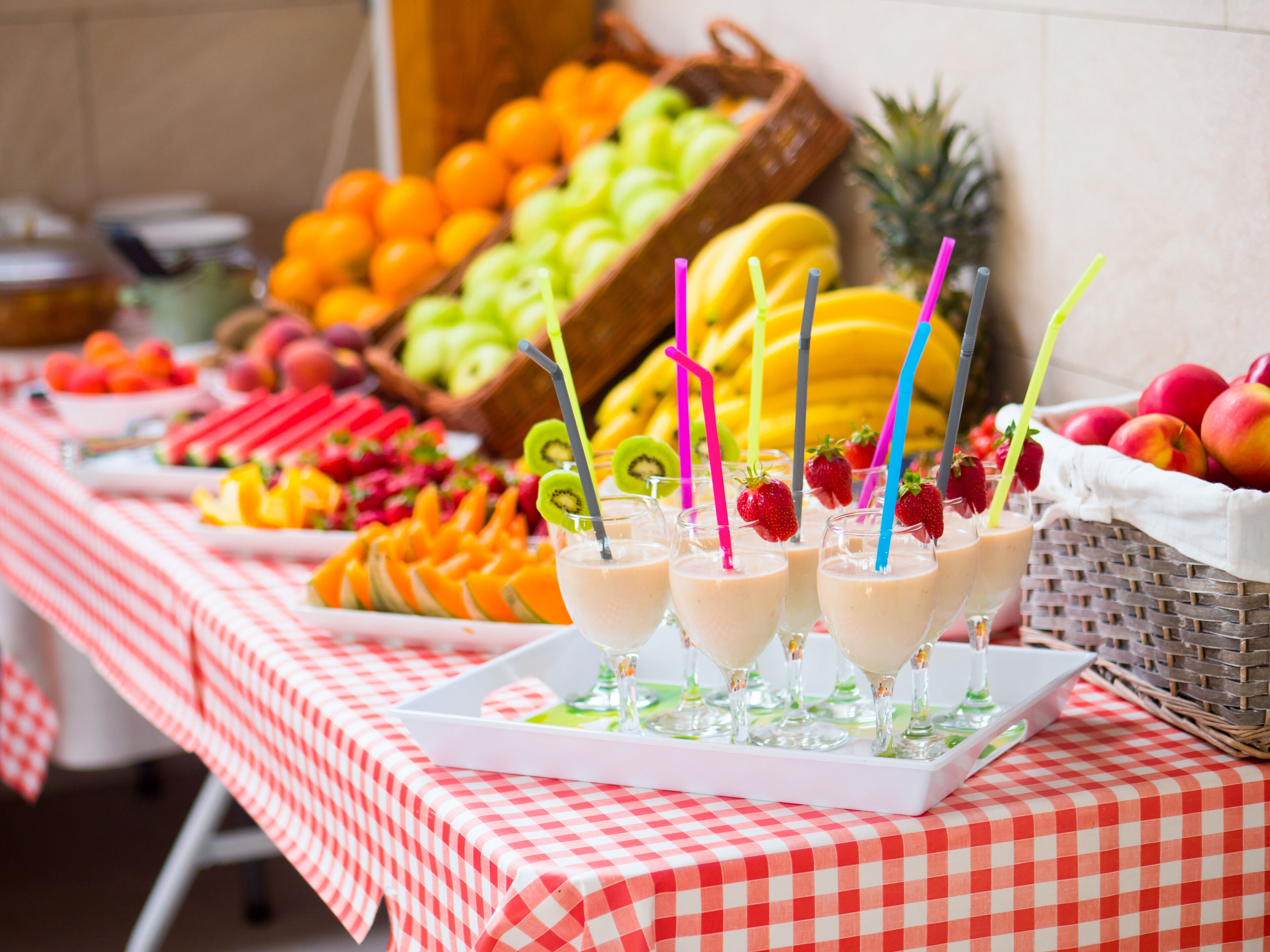 Un tavolo con tovaglia a quadretti rossi e bianchi, pieno di frutta varia, un vassoio di smoothie e un cesto di mele.