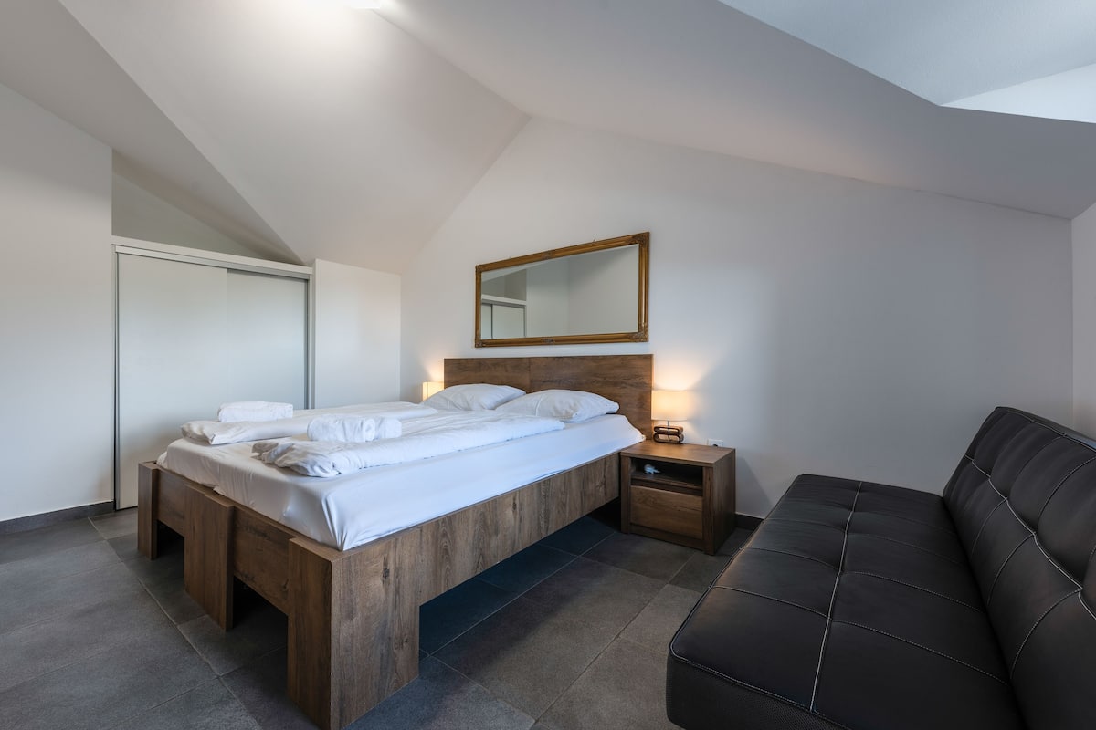 A modern bedroom with a wooden bed, a nightstand, a black leather sofa, and a mirror on the wall.
