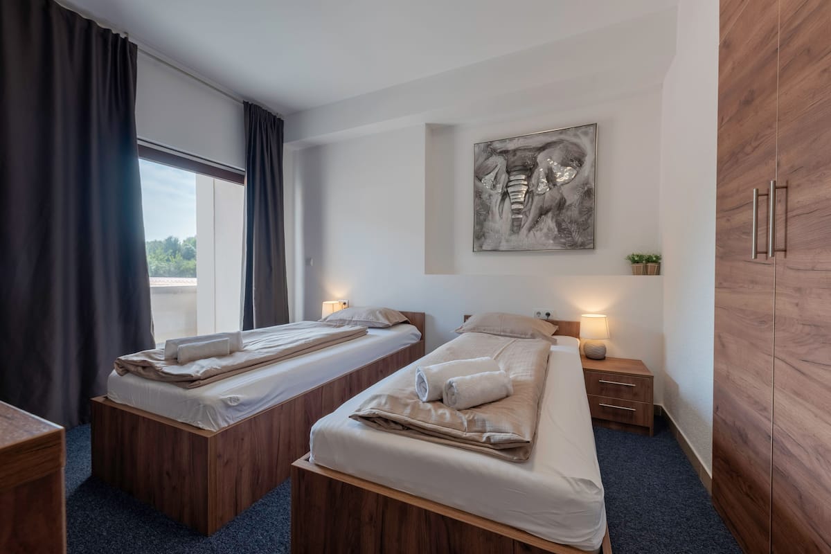 A hotel room with two wooden beds, a large window with curtains, a wall-mounted artwork, and a wardrobe.