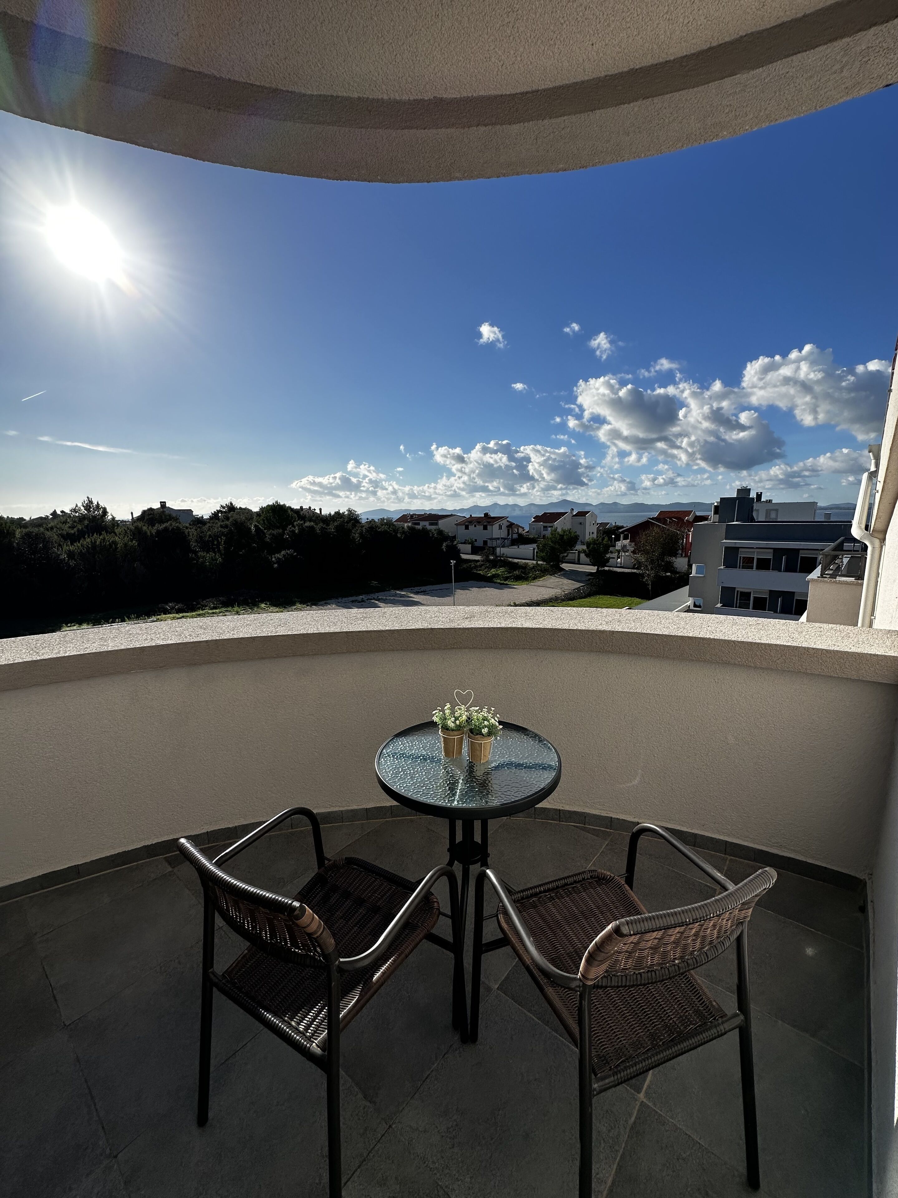 A balcony with a round glass table and two wicker chairs, overlooking a residential area with houses and mountains in the distance.