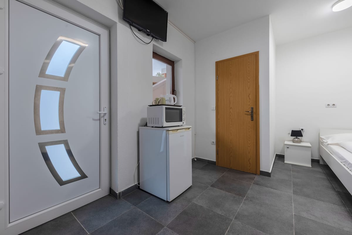 A compact hotel room with a white door, a small kitchenette with a microwave, a wooden door, a bedside table with a lamp, and a bed.