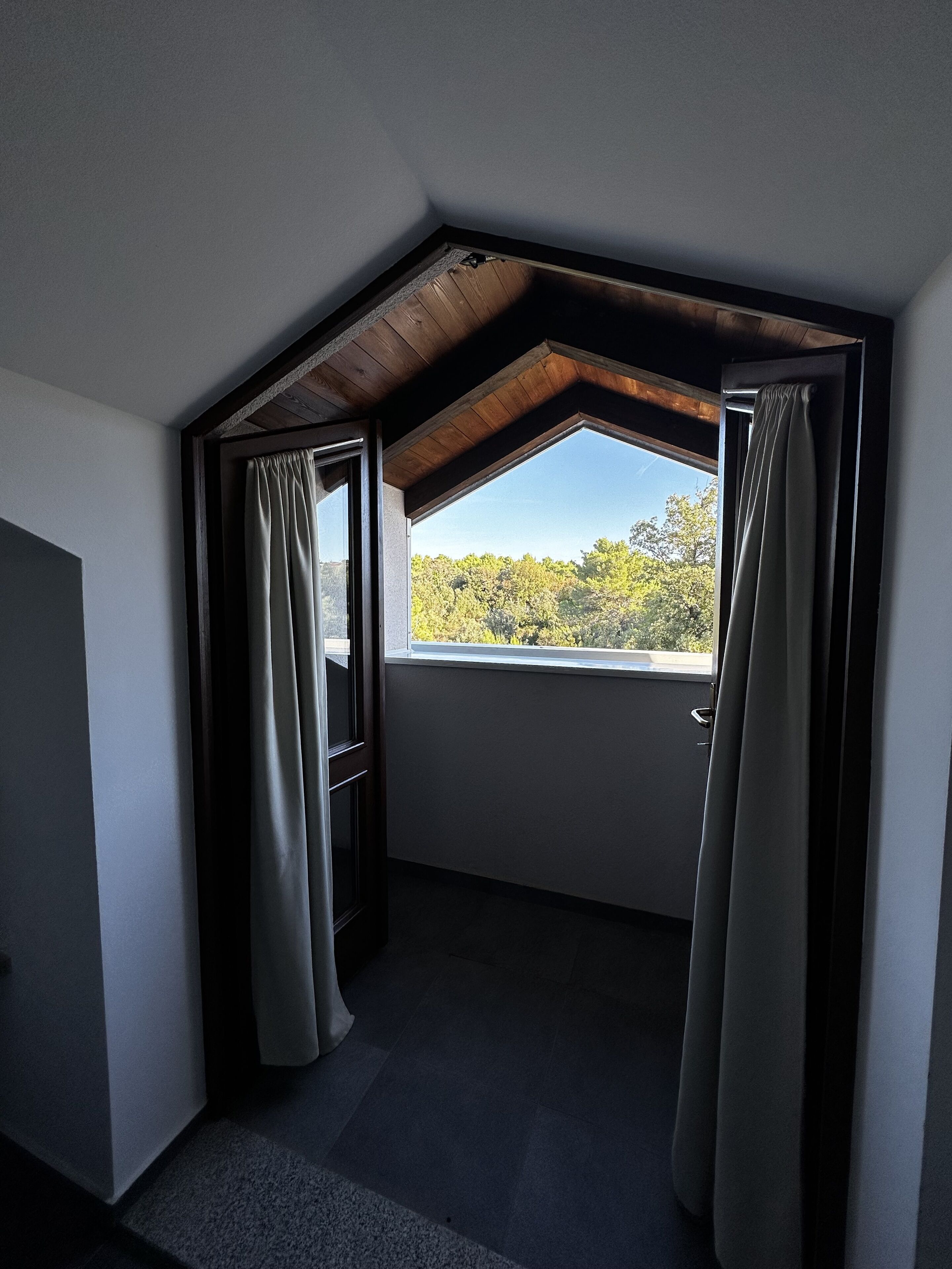 A room with a sloped ceiling, a large window with curtains, and a view of trees outside.