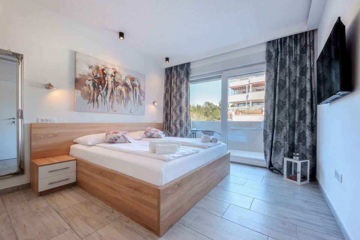 A modern bedroom with a wooden bed, a large painting, a balcony with curtains, and a TV mounted on the wall.