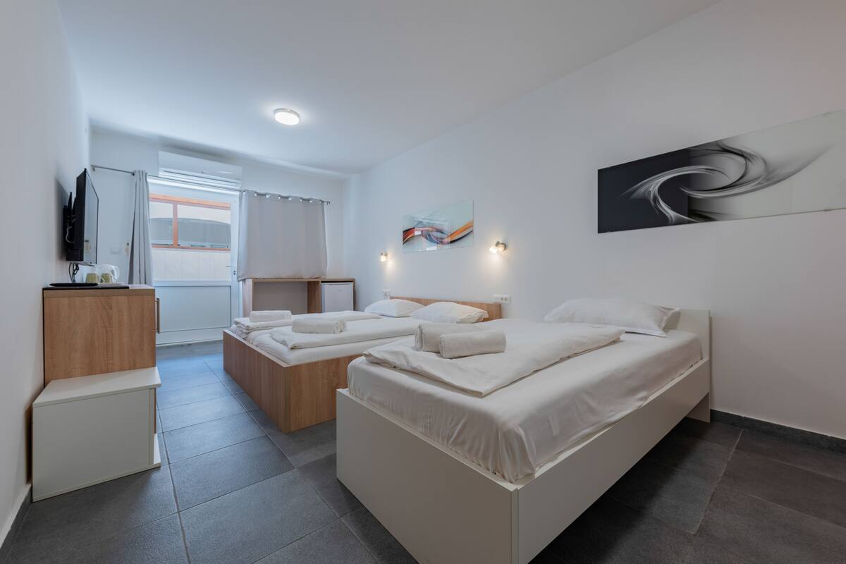 A modern hotel room with two single beds, a small desk, a television, and a view of the outdoors.