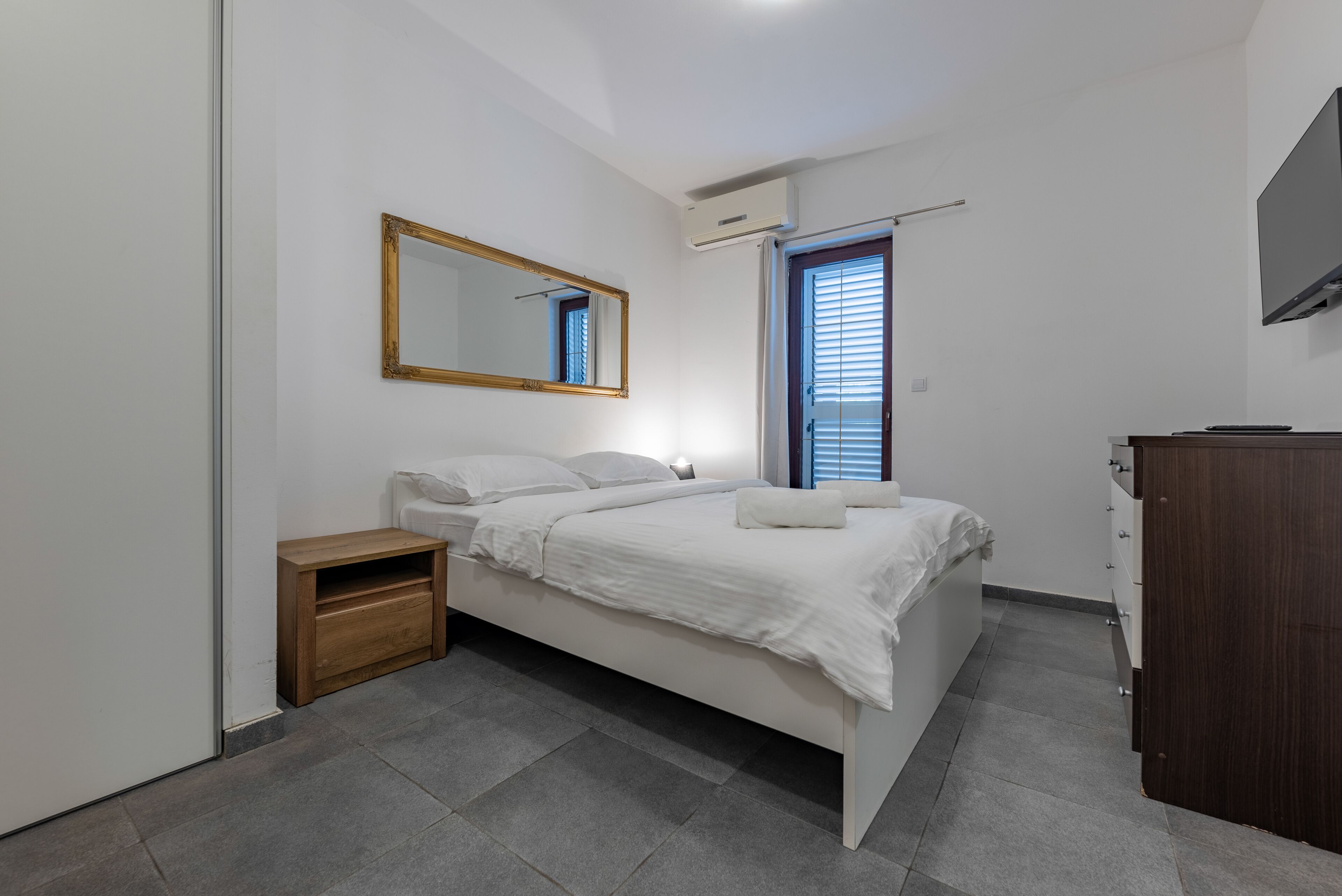 Quarto com cama, mesa de cabeceira de madeira, espelho, janela com persianas e televisão na parede.