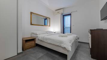 Quarto com cama, mesa de cabeceira de madeira, espelho, janela com persianas e televisão na parede.