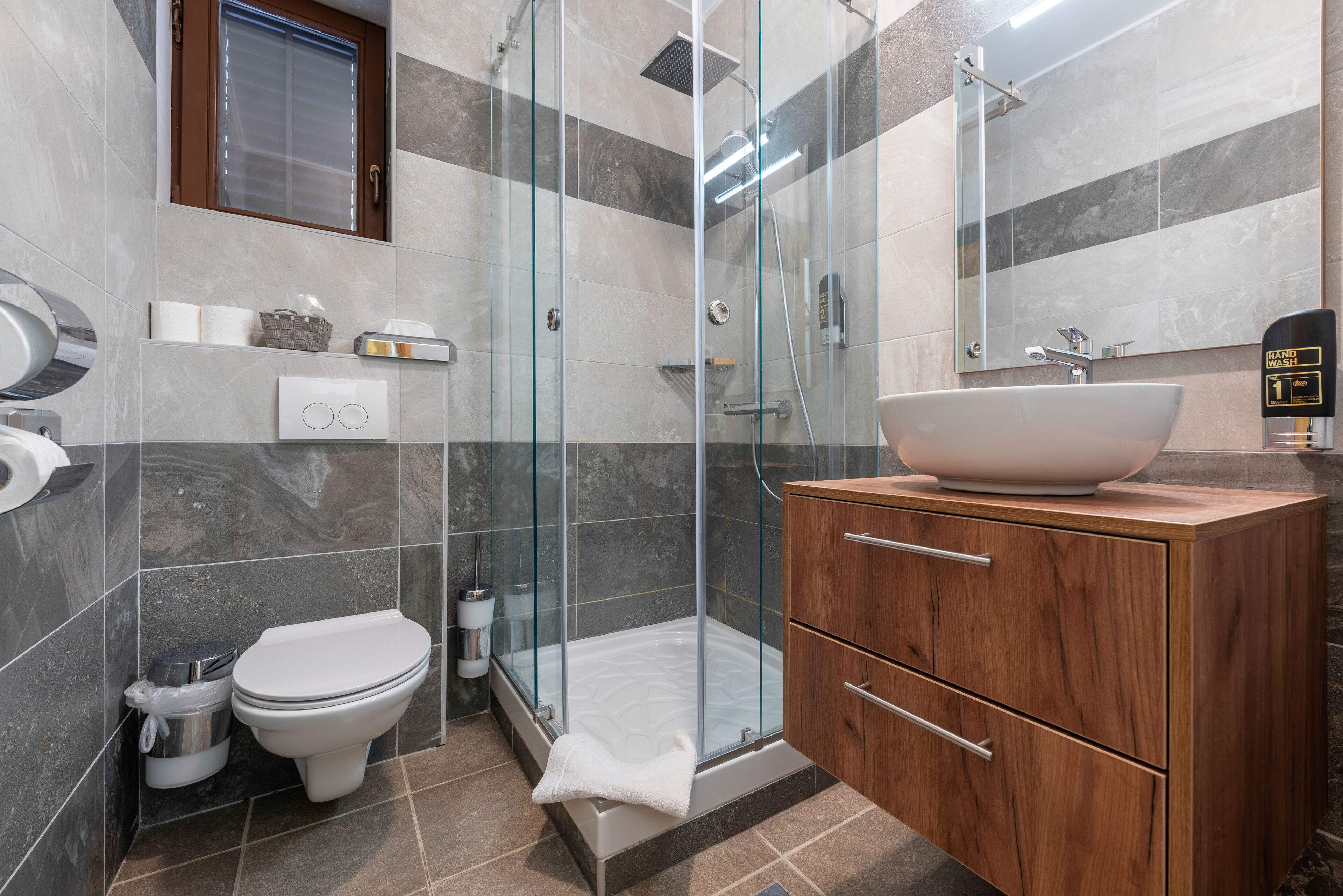 Un baño moderno con ducha de vidrio, tocador de madera con un lavamanos grande y un inodoro.