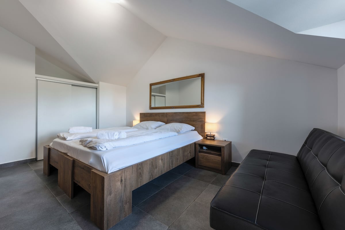 A modern bedroom with a wooden bed, a nightstand, a black leather sofa, and a mirror on the wall.
