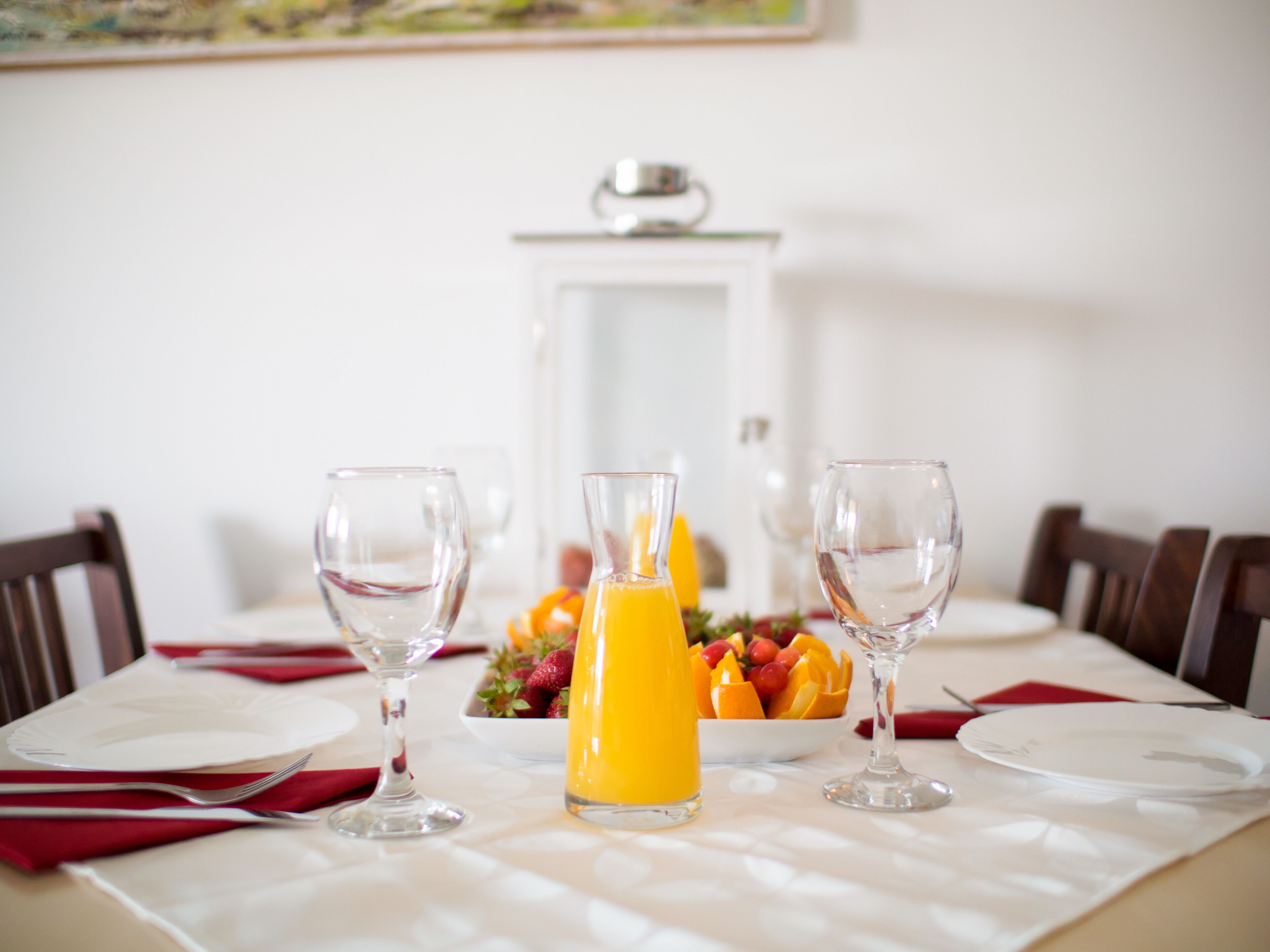 A dining table set with white plates, red napkins, and clear wine glasses. In the center, there is a pitcher of orange juice and a bowl of fruit.
