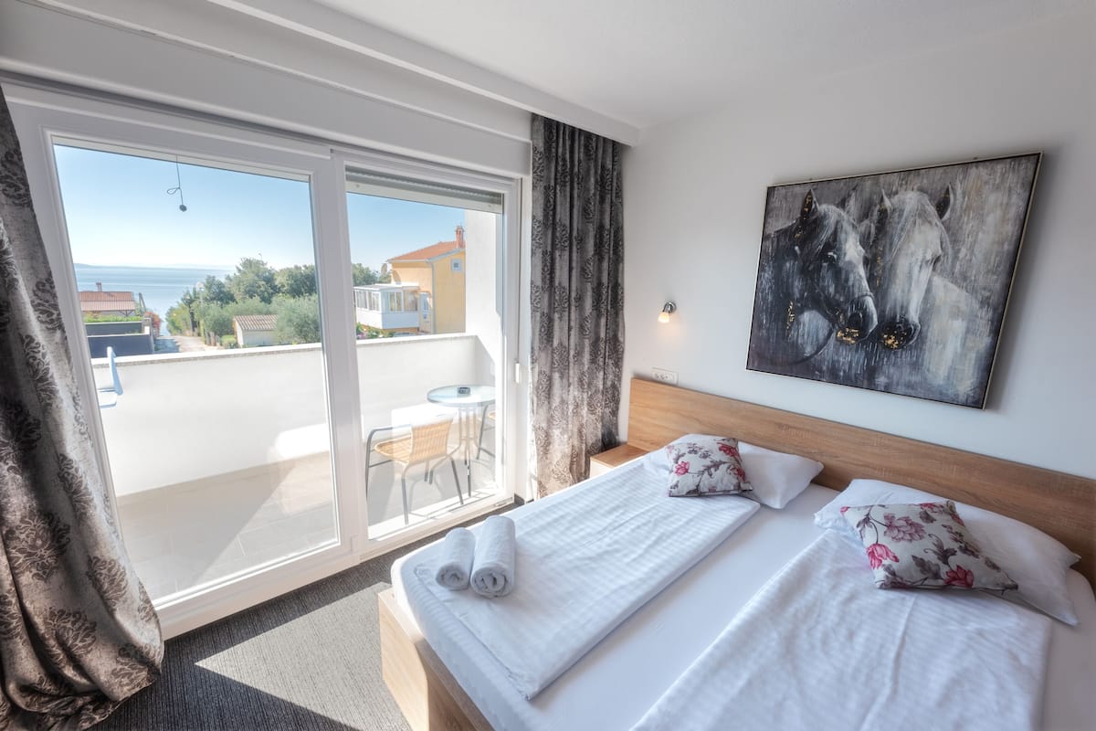 A hotel room with a balcony, a bed with white linens, a painting of horses, and a view of the sea.