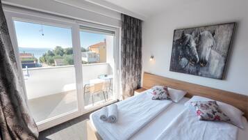 Habitación de hotel con balcón, cama con ropa blanca, pintura de caballos y vista al mar.