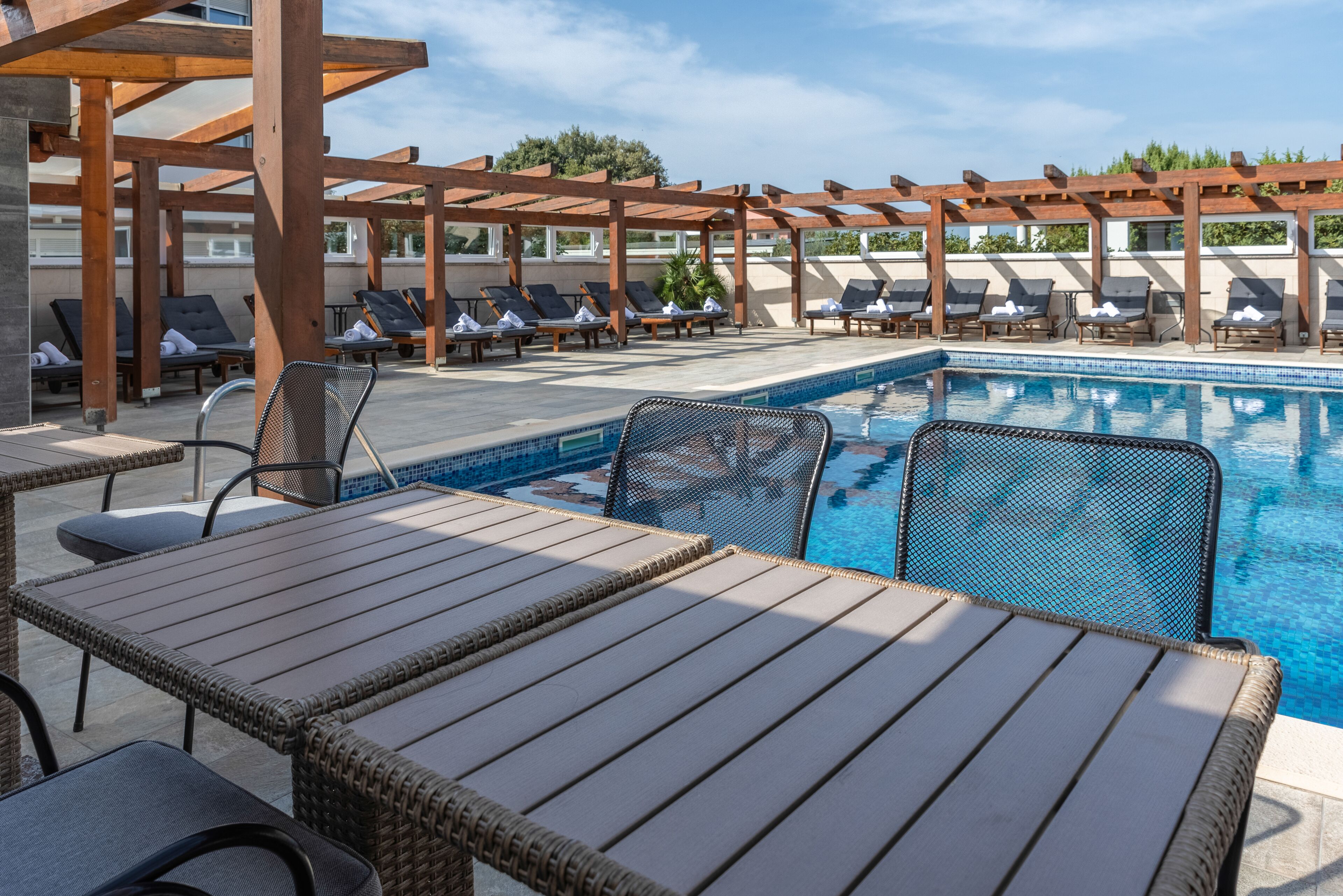 Un área junto a la piscina con sillones, mesas y una pérgola.